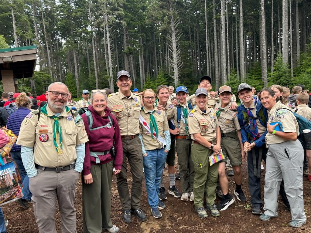 Group of scouts and leaders posing for a photo in a wooded area, wearing scout uniforms and various badges.