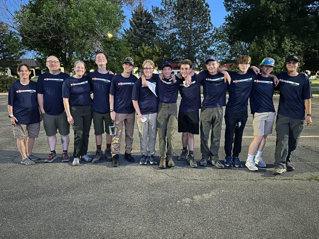 A group of twelve people standing in a line, all wearing matching navy blue shirts, in a parking lot with trees in the background. They are smiling and posing together.