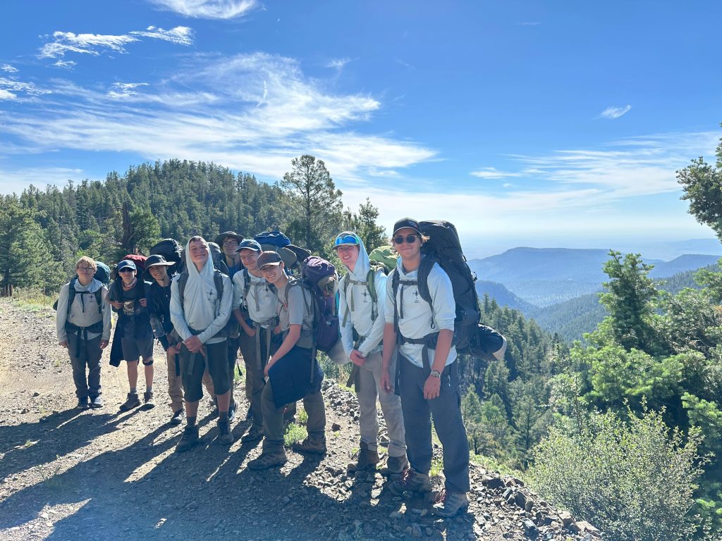 A group of hikers posing for a photo on a mountain trail with lush trees and a scenic valley view in the background.