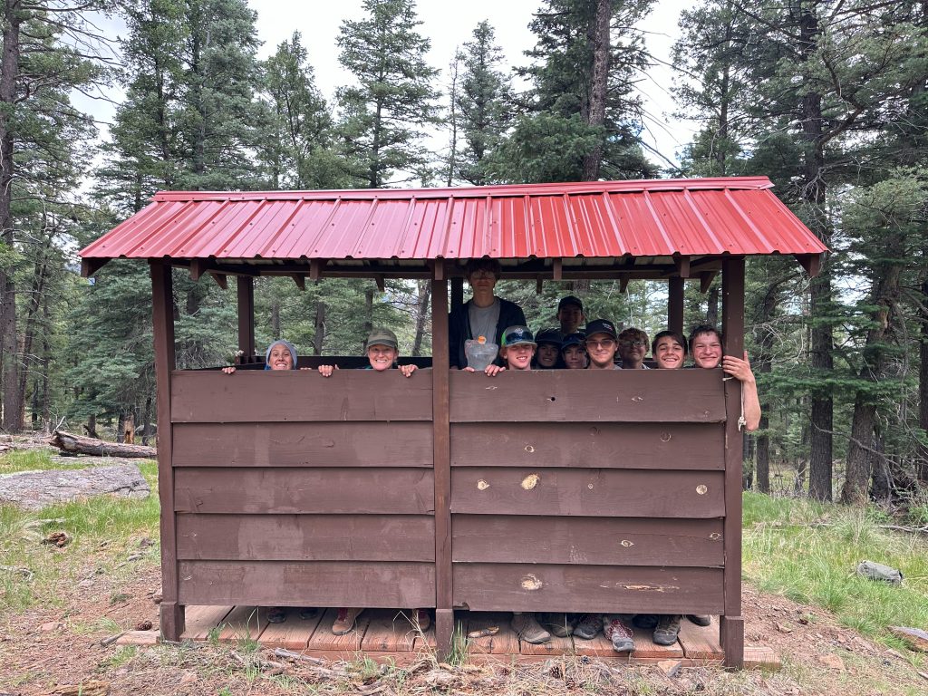A group of people smiling and posing together inside a wooden shelter with a red metal roof, surrounded by tall trees in a forested area.