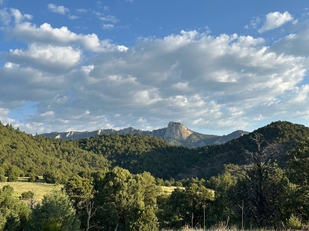 A scenic view of rolling green hills and majestic mountain peaks under a partly cloudy blue sky.