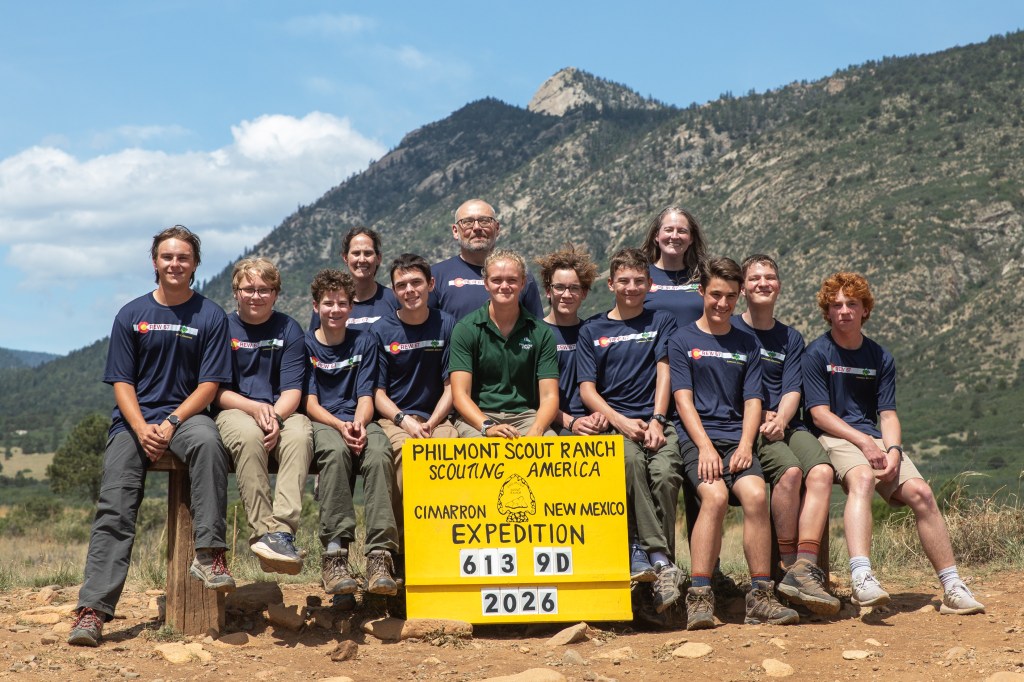 A group of twelve individuals posing for a photo in front of a mountainous landscape, wearing matching blue shirts and khaki shorts. They are gathered around a sign that reads 'Philmont Scout Ranch, Scouting America, Cimarron, New Mexico, Expedition 613 9D 2026.'