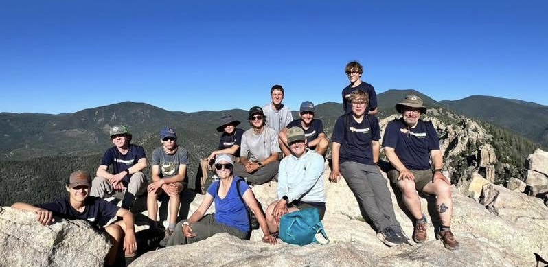 A group of eleven people sitting on a rocky outcrop at the top of a mountain, surrounded by green hills and a clear blue sky.